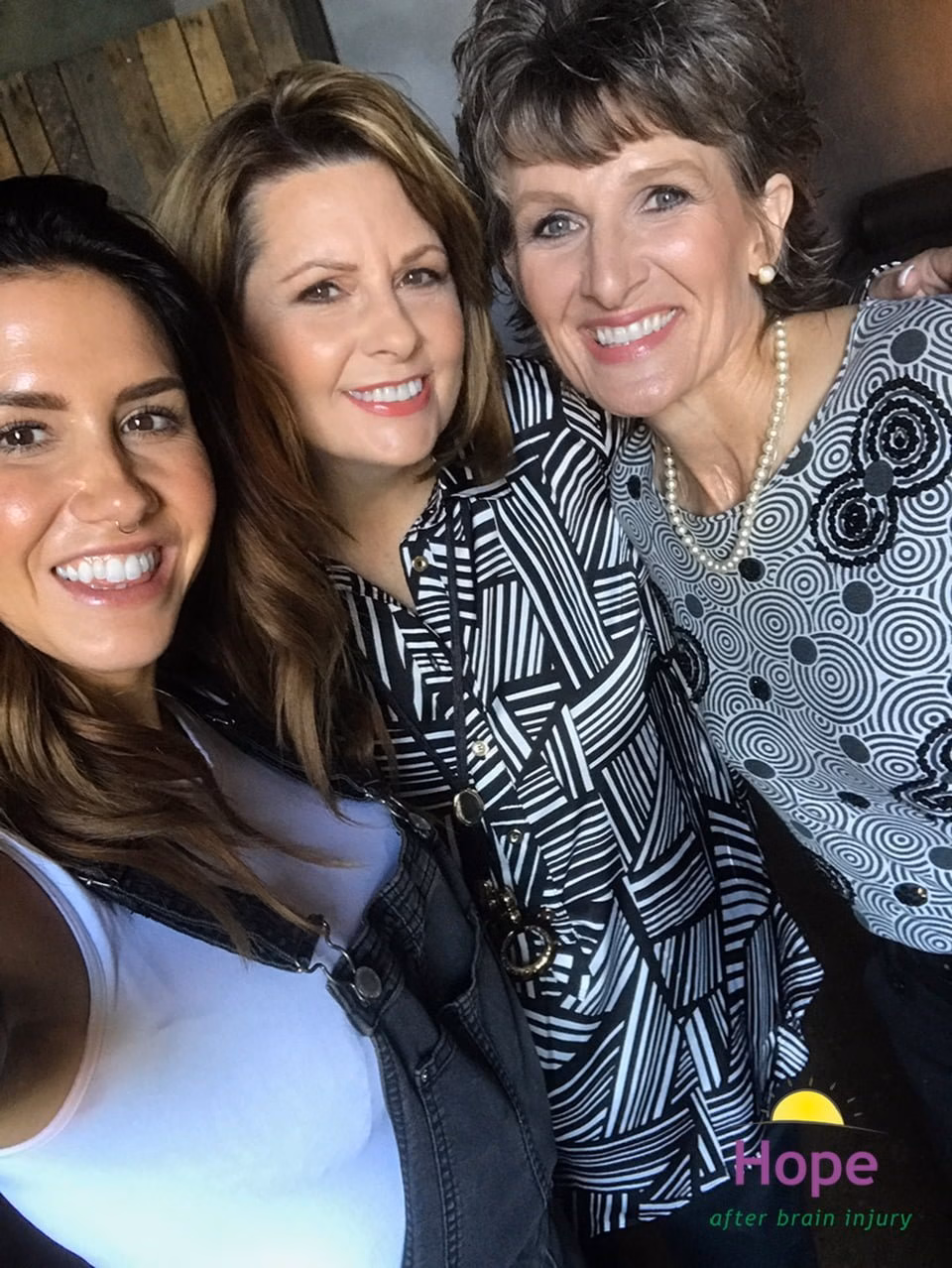 Three smiling women pose closely together for a selfie. The woman on the left wears a white top and overalls, the two on the right wear patterned tops. In the bottom right corner, theres a Hope after brain injury logo.